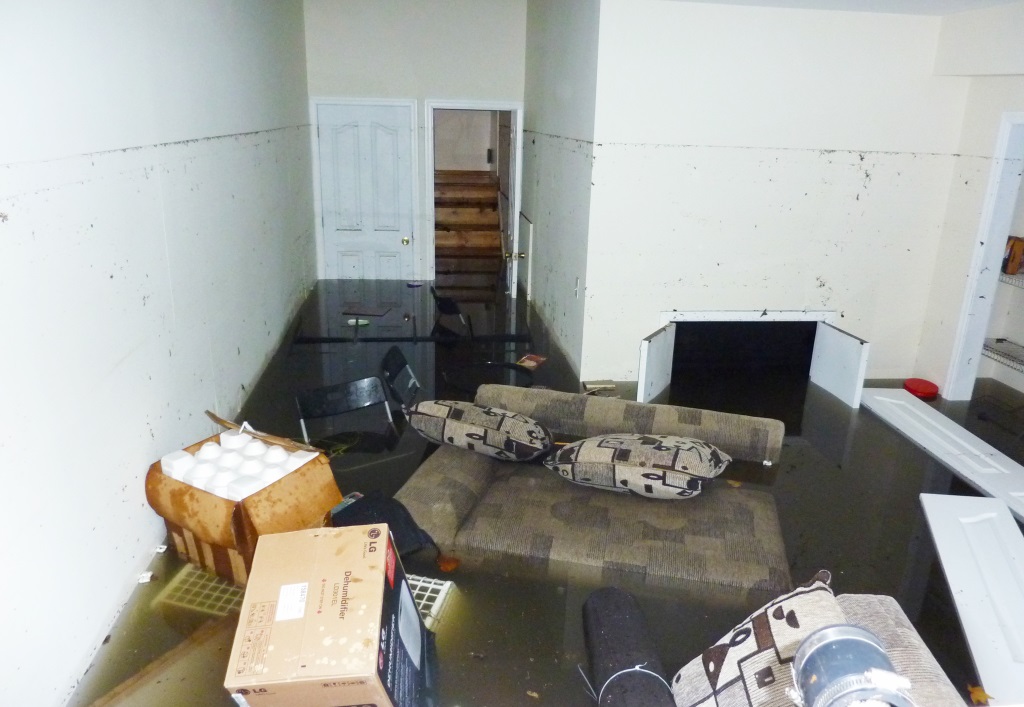A flooded basement living area with dark water covering the floor, submerging furniture including a sectional sofa, boxes, and other belongings, with water damage visible on the white walls.