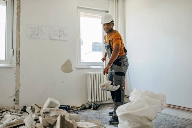 A construction worker in overalls and an orange shirt is renovating a room, standing near a radiator amid debris and plastic sheeting scattered on the floor.