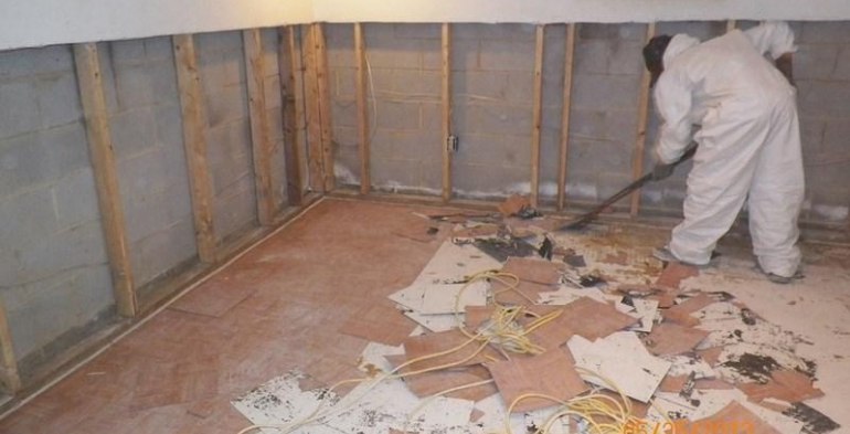A person in white protective gear is working in a basement or lower level room with exposed wooden wall framing, concrete block walls, and a floor covered with debris including damaged materials, papers, and what appears to be flood or water damage cleanup materials.RetryClaude can make mistakes. Please double-check responses.