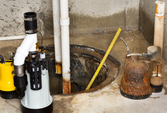 A basement sump pump pit showing water damage repair work with a new white sump pump on the left, a measuring stick in the concrete pit, and a rusty old pump on the right.